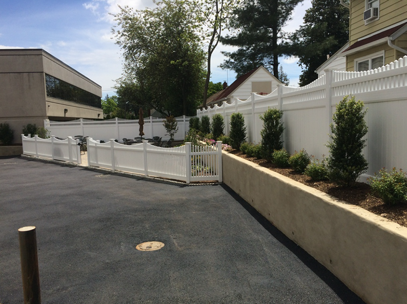 White picket fence and landscaping along a paved area, with a building in the background.