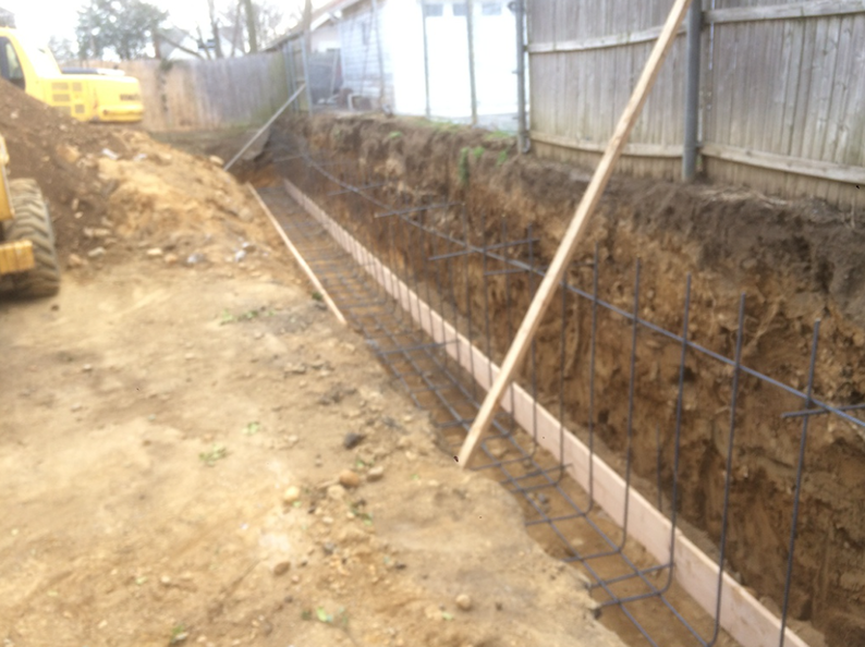 Trench dug, wooden formwork and rebar in place, next to a fence, preparing for concrete foundation.