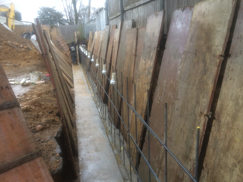 Construction trench with wooden shoring, metal reinforcement, and concrete base along a fence line.