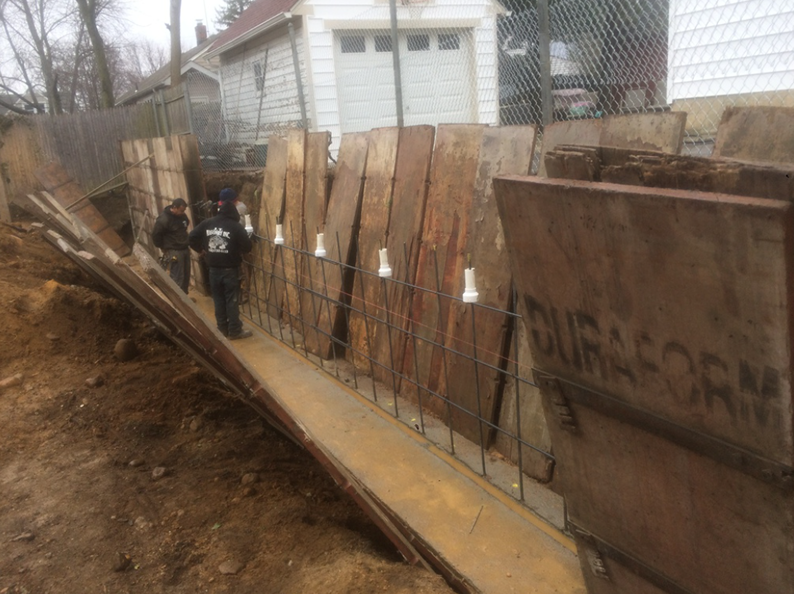 Construction workers near a trench with wooden and metal forms. Earth and fence are visible.