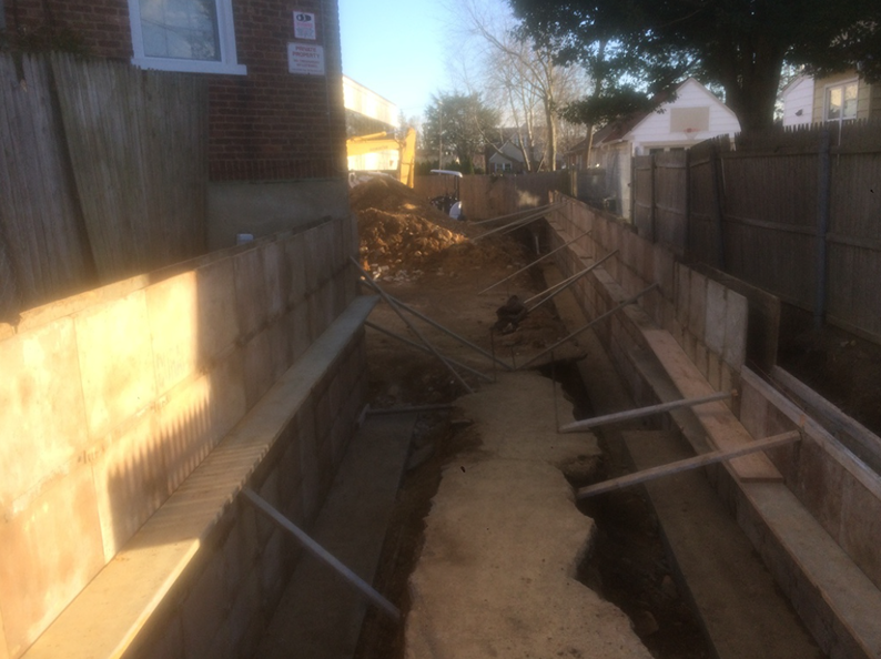 Construction site with deep trench, wooden retaining walls, and backhoe in the background.
