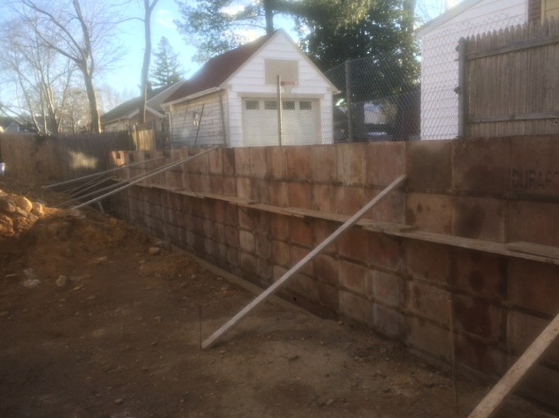 A brown retaining wall under construction with wooden braces, set in a residential backyard.
