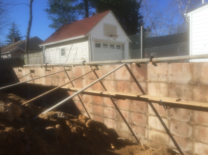 Red-roofed garage behind a retaining wall under construction. Wooden supports, brick blocks, and dirt are visible. Sunny day.