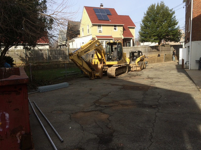 Yellow excavator and skid steer in a paved yard, house with solar panels in the background.