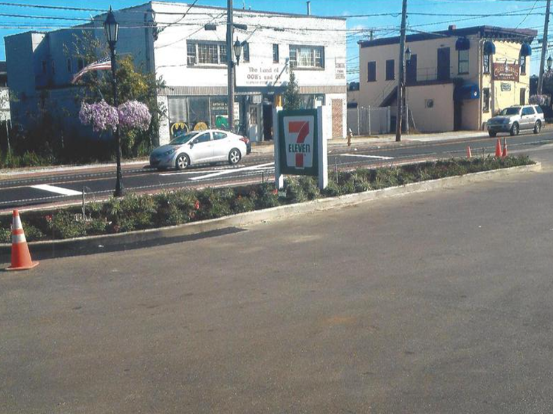 Street view of a 7-Eleven, car driving by, landscaping, buildings in background.