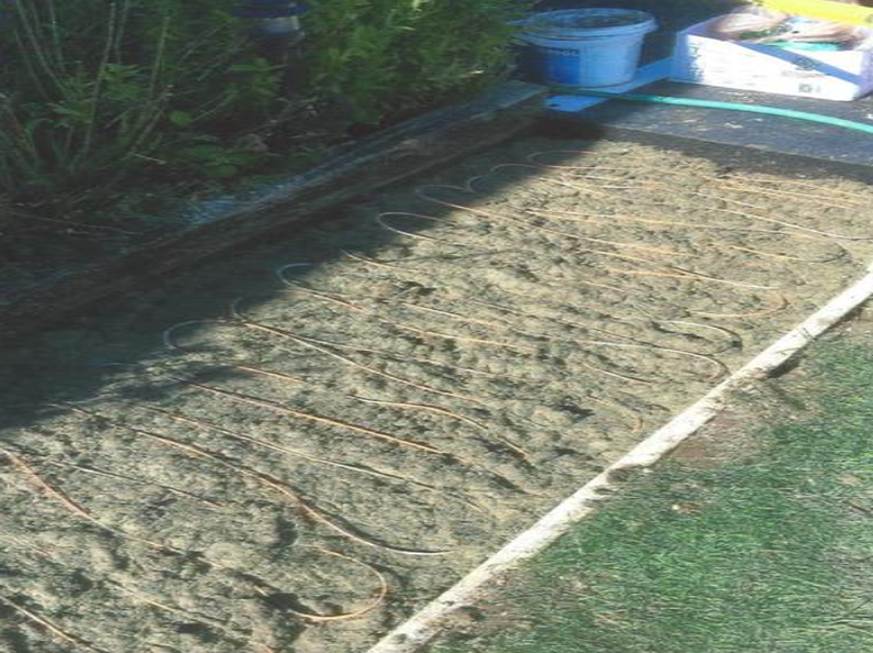 Copper pipes laid on a sandy surface for outdoor heating, next to grass and garden.