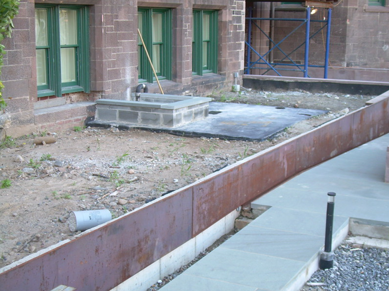 Exterior construction site with a rusted metal barrier, concrete path, and brick building with green-framed windows.