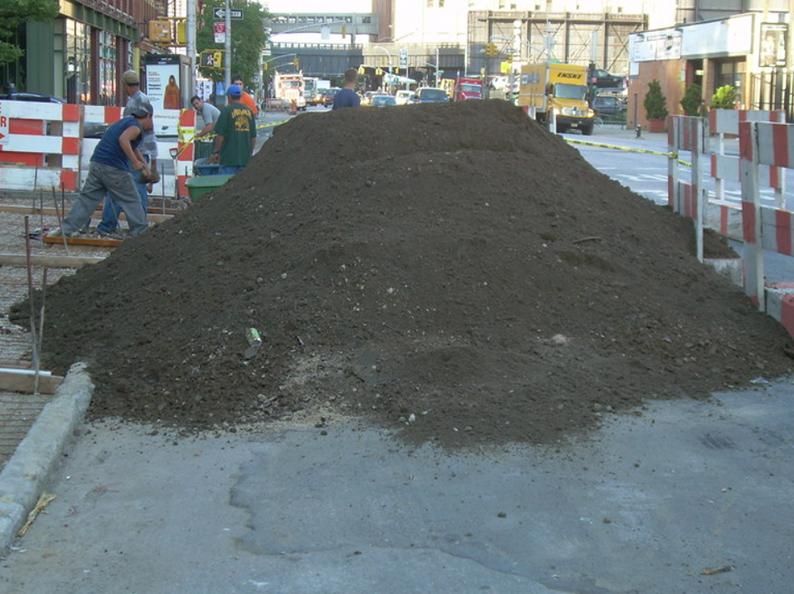 Large pile of dark dirt on a city street with construction workers in the background.