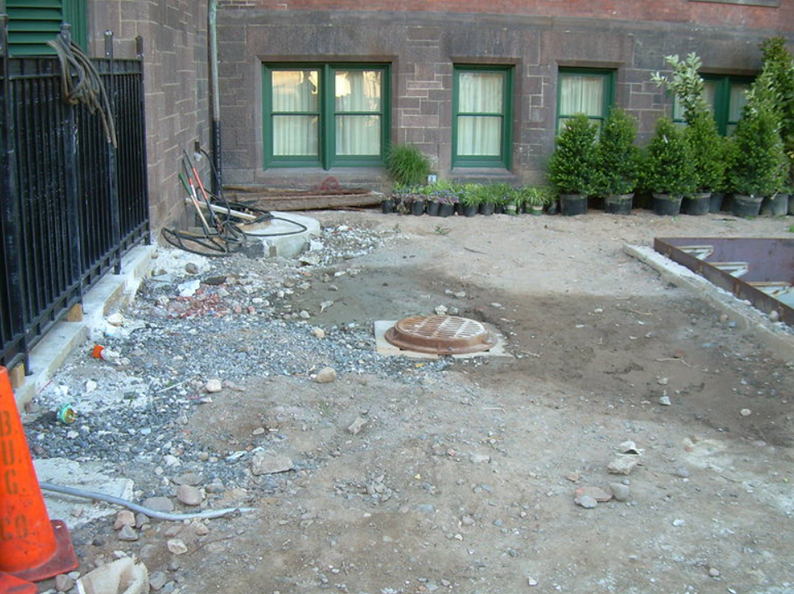 Construction site with debris, manhole cover, and potted plants next to a brick building.