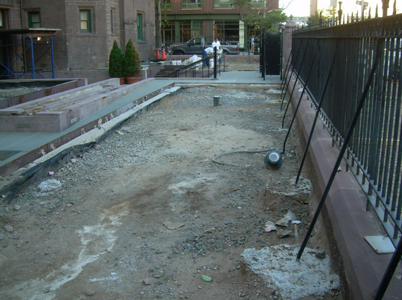 Construction site: bare earth between a concrete planter and black metal fence, with building in background.