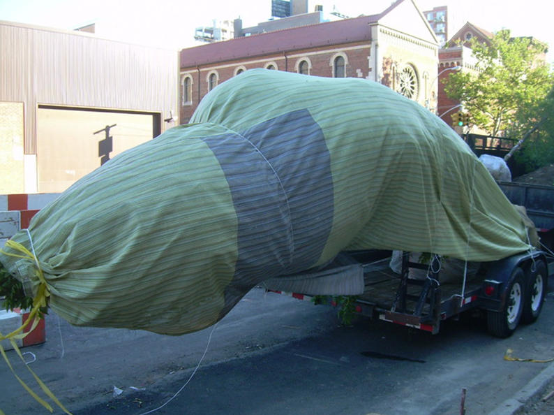 A large object covered in green cloth is on a trailer, in a city setting.