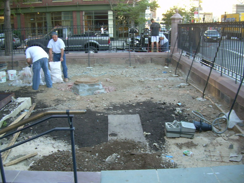 Workers filling in a dirt area near a wrought-iron fence, with a truck, and building in the background.