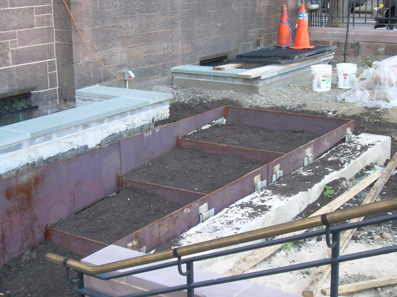 Metal steps under construction, filled with dirt. Construction site with brick wall, handrail, and cones.