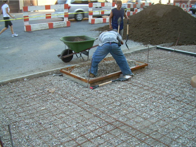 Construction workers paving a road. One shovels gravel within a frame, others work nearby with a wheelbarrow and a pile of dirt.
