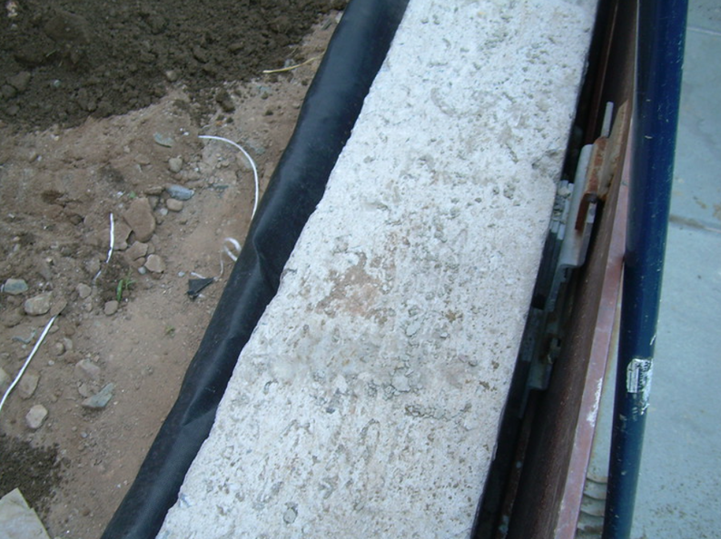 Concrete block next to black landscaping fabric. Dirt and a blue pole are visible.