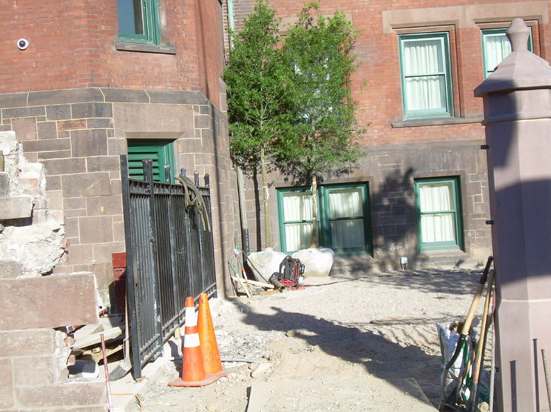 Construction site with brick buildings, metal fence, gravel, and orange traffic cones.