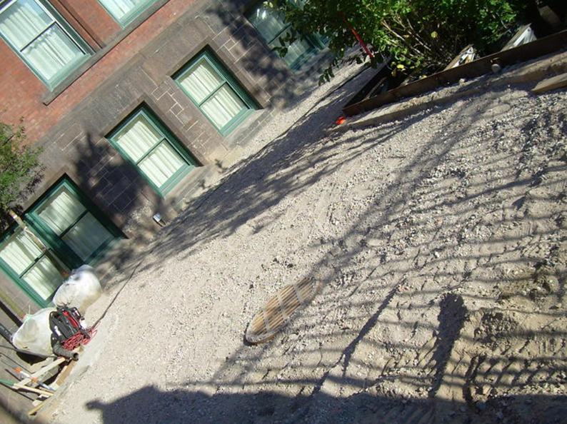 Exterior shot of a building with green window frames and a gravelly surface. A shadow from a fence stretches across the ground.