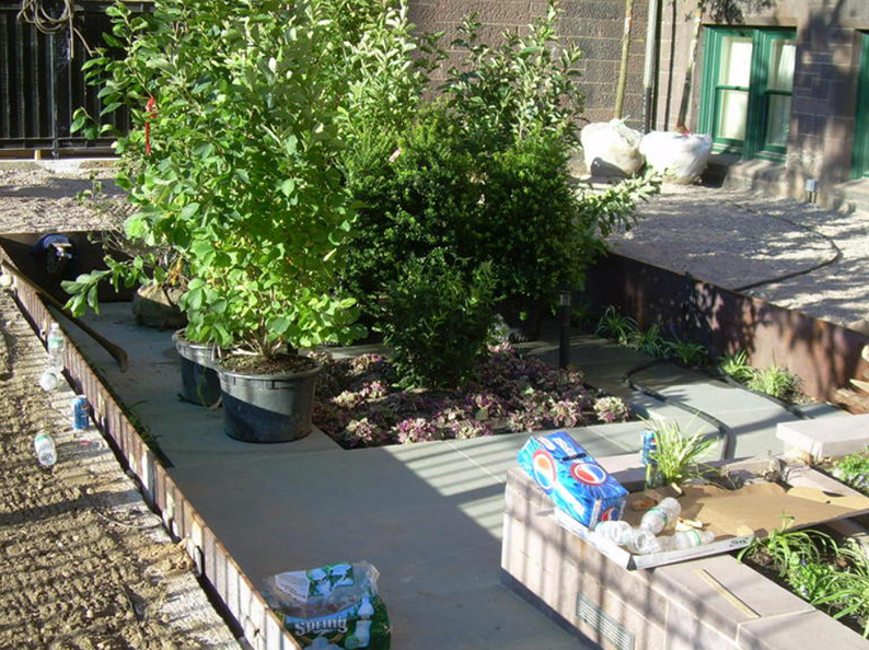 A garden with potted plants, brick path, and a concrete walkway next to a metal border.
