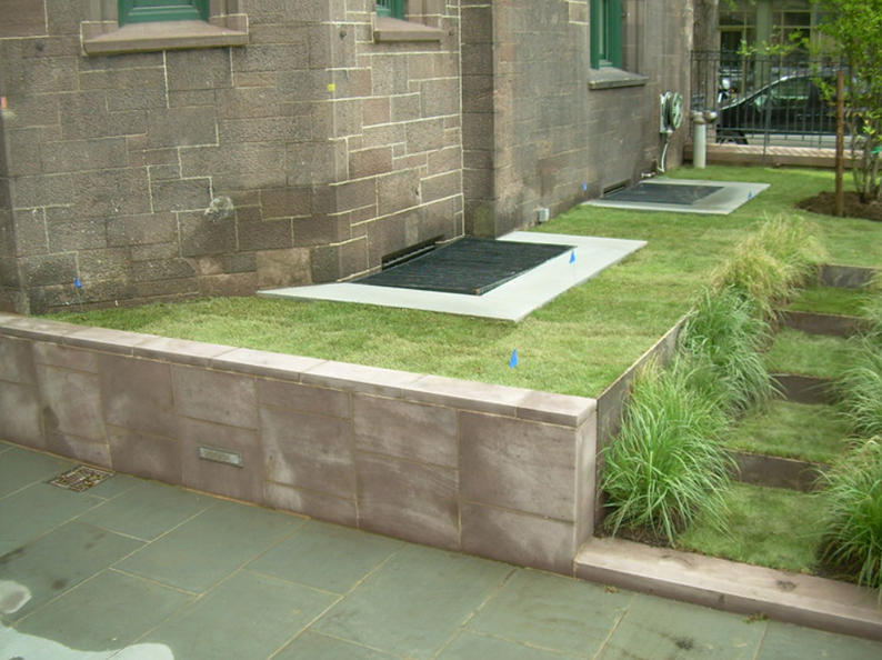 Low retaining wall with grass and rectangular grates in the ground alongside a stone building.