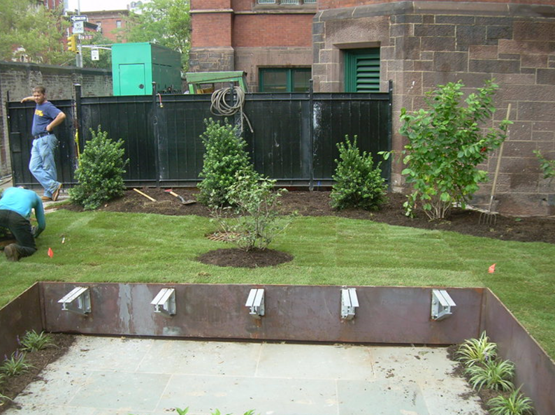 Workers installing landscaping with a dark fence and a brick building in the background.
