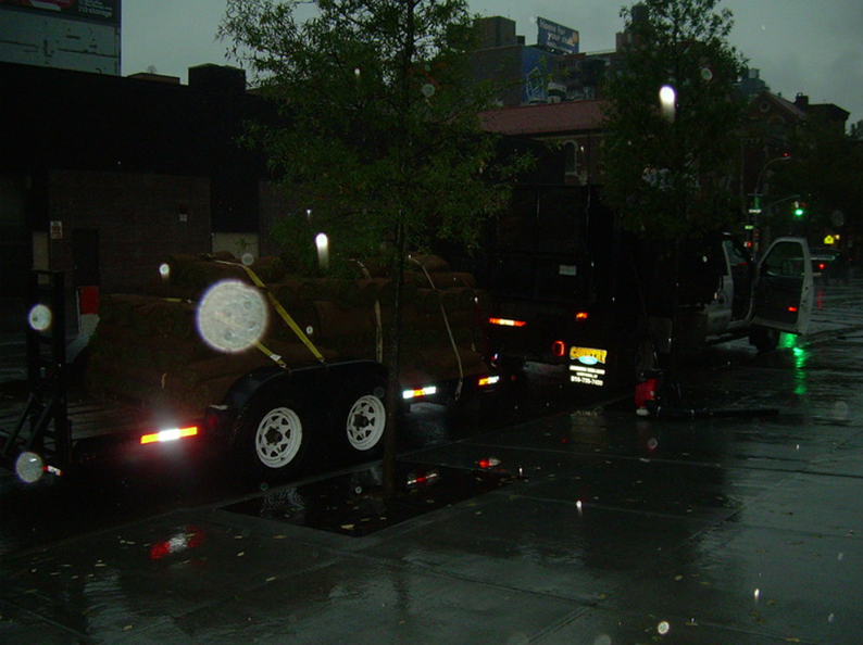 Rainy street scene: Trailer with wood, lights, and open van, reflecting in puddles.