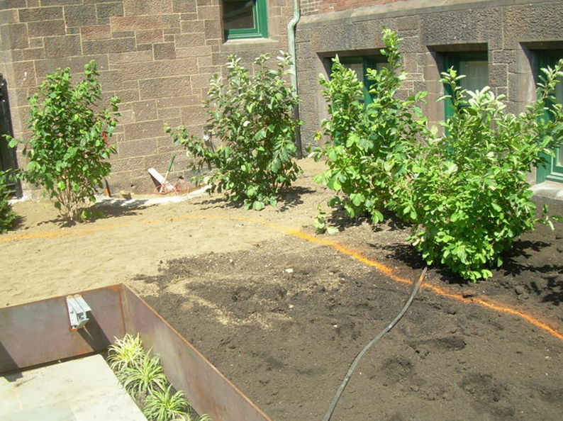 A garden bed with bushes, fresh soil, and a metal planter in front of a brick building.