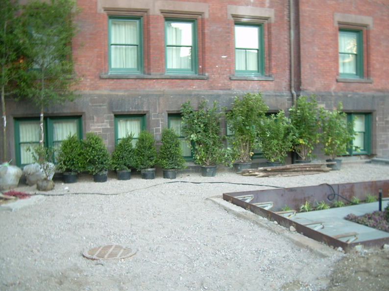 A brick building with green-trimmed windows and potted green bushes along a gravel-covered ground.