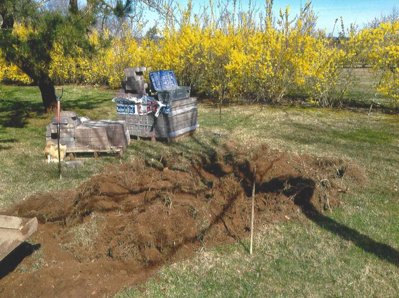 A patch of dug-up earth in a yard, with a container of supplies in the background near a flowering bush.