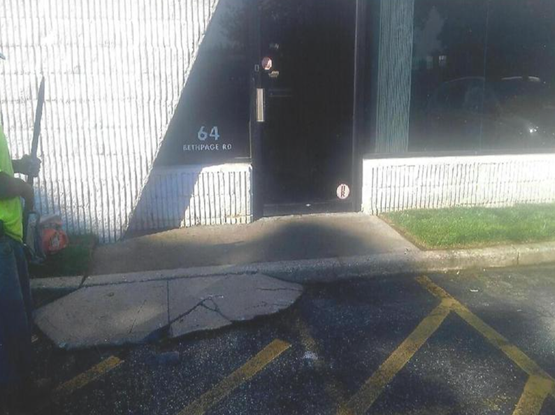 Cracked concrete on sidewalk in front of a building, marked parking space with faded yellow lines, side view.