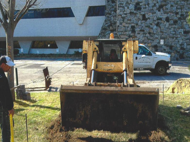 Construction worker watches a skid steer loader with dirt in its bucket, in front of a building, and a white truck.