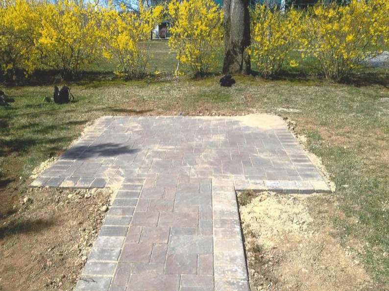 Brick pathway leading to a square patio, surrounded by dirt and grass, with yellow flowering bushes in the background.