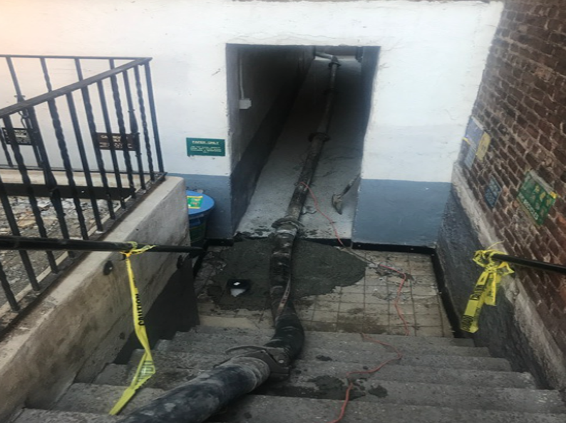 Staircase blocked by concrete hose. Entrance with blue/white walls. Brick wall on right. Yellow caution tape.