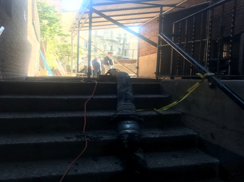 Concrete stairs lead up. A dark hose lies on steps. Two people at the top under scaffolding.