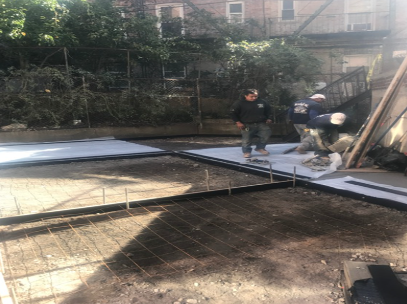 Workers laying concrete foundation in a backyard; rebar frame visible, surrounded by trees and buildings.