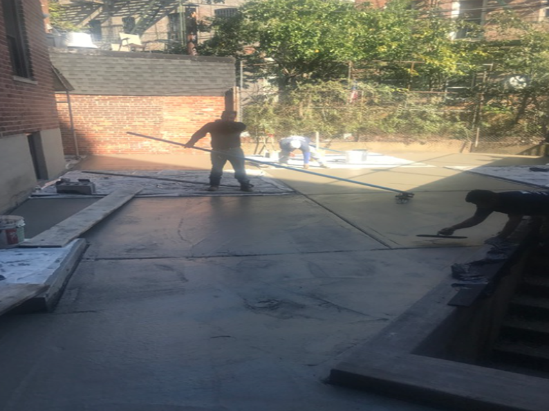Construction workers smoothing wet concrete on a patio, surrounded by brick buildings and trees.