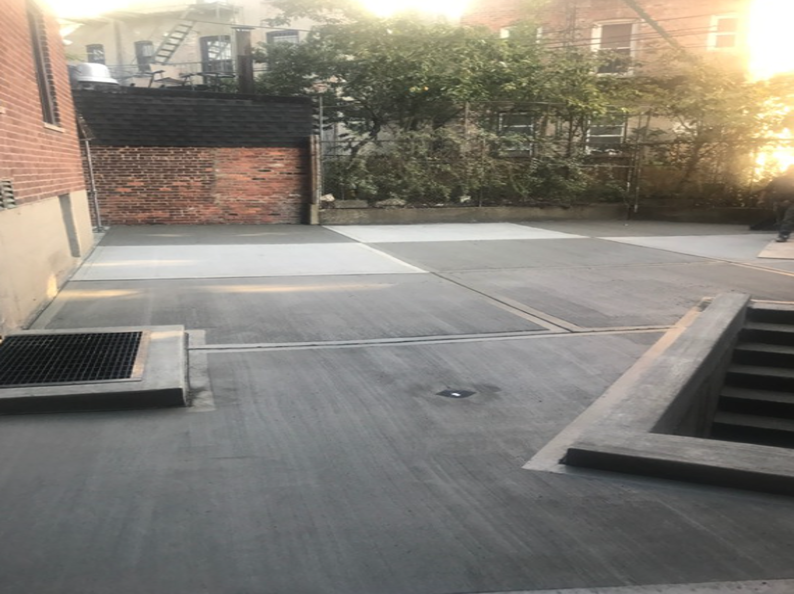 Concrete courtyard with a grated drain, brick wall, and stairs. Trees and buildings in the background.