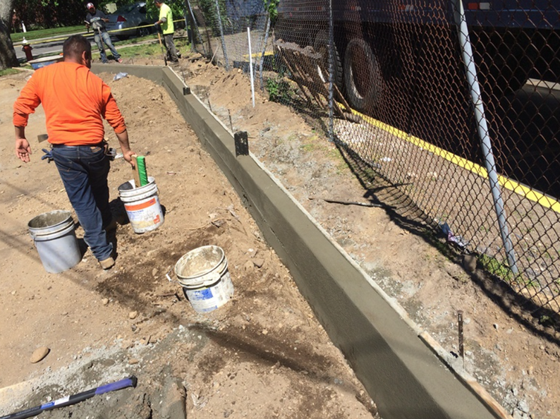 Construction worker pouring concrete curb next to a chain-link fence. Buckets and tools nearby.