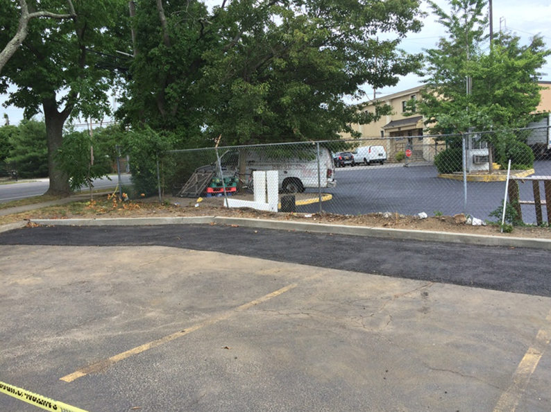 An asphalt parking lot with a chain-link fence, van, and trees; commercial buildings in the background.