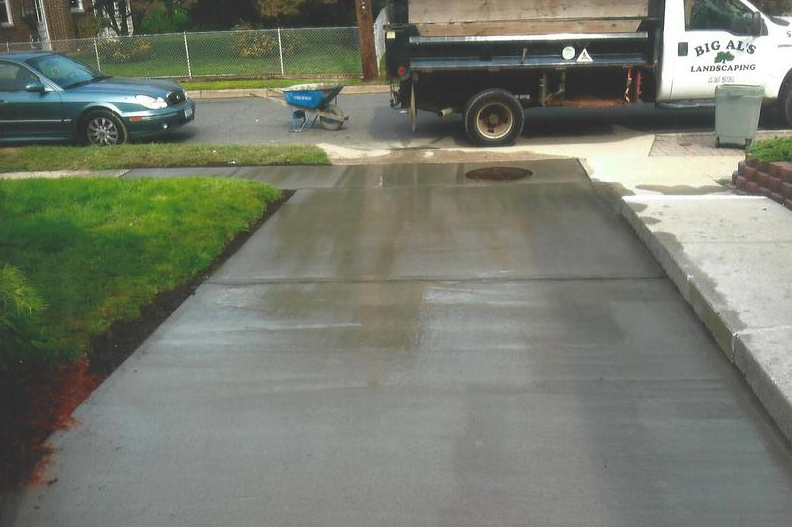 Freshly poured concrete driveway; green lawn, car, landscaping truck, wheelbarrow.