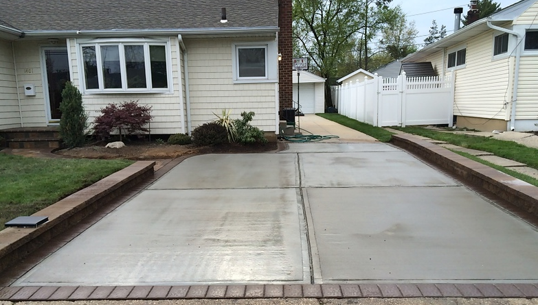 Newly poured concrete driveway with brick borders next to a light-colored house with a bay window.