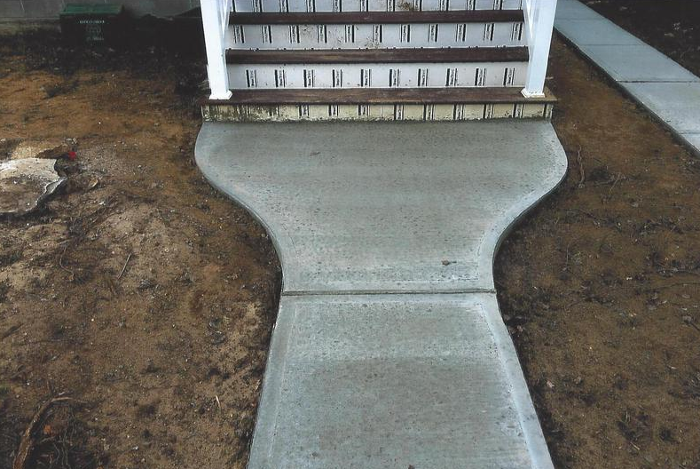 Concrete sidewalk leading to wooden stairs of a building; surrounded by dirt and a parallel sidewalk.