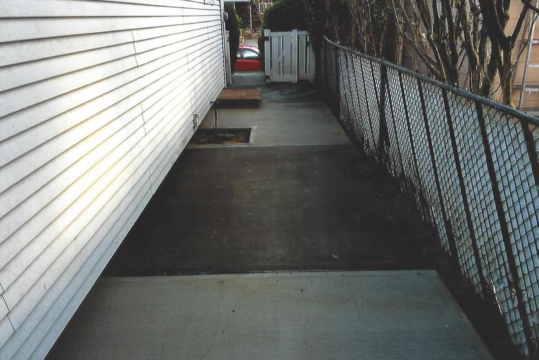 Concrete walkway along a white-sided building and a chain-link fence. Small opening to the right, leading to a red car.