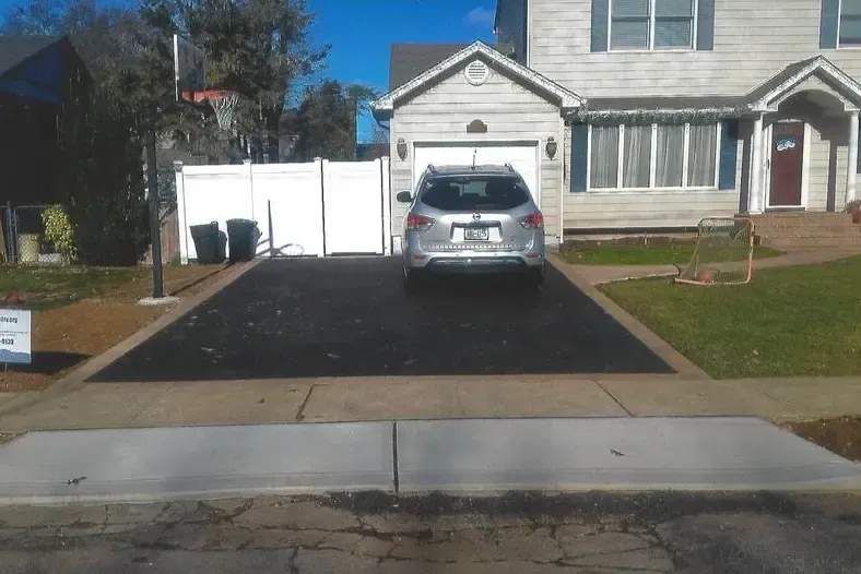 Black paved driveway in front of a two-story house with a car parked in the garage, a white fence, and a blue sky.