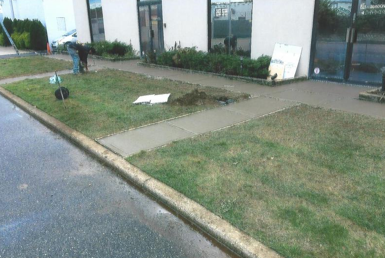 Exterior view of a building with fresh concrete sidewalk and patches of grass. A person works nearby.