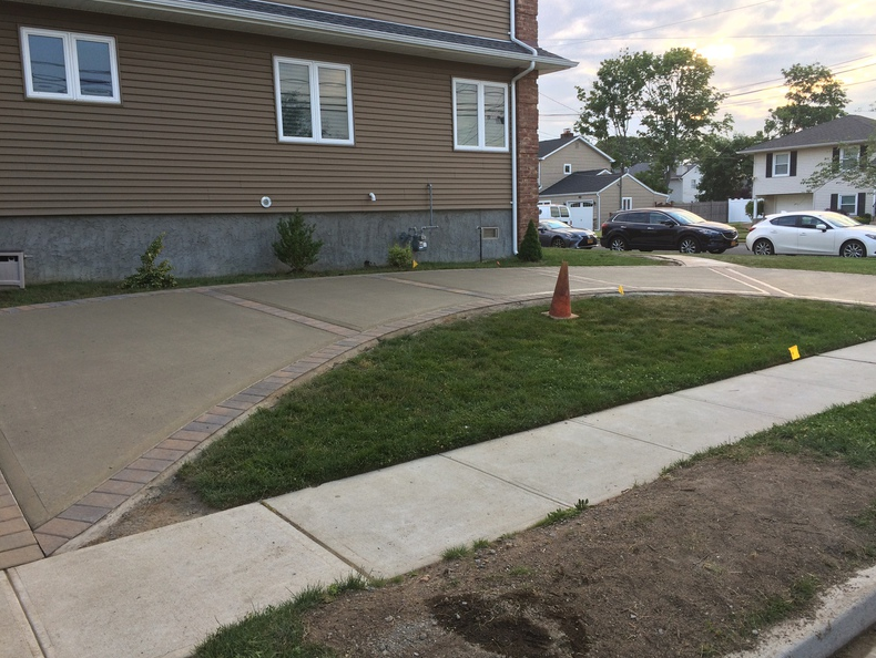 A concrete driveway curves past a small lawn in front of a two-story beige house; a sidewalk is in the foreground.