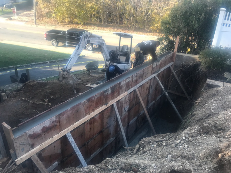Construction site: workers pouring concrete into wooden forms for a retaining wall. A backhoe and truck are present.