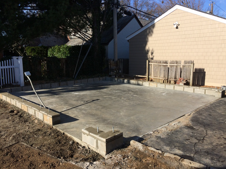 Concrete patio construction in a backyard, surrounded by brick, with a garage and fence visible.