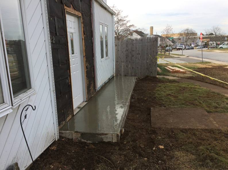 Newly poured concrete porch and entryway, partially built, next to a white house with a fence.