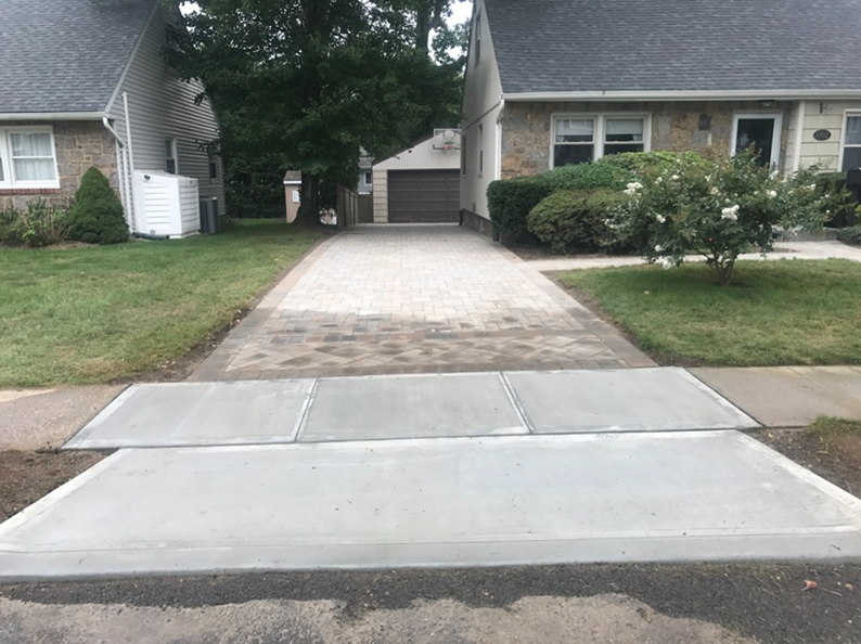 Driveway with brick pavers leading to a garage between two houses; new concrete sidewalk in front.
