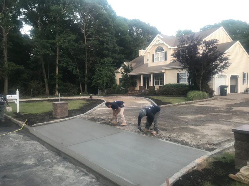 Two workers smoothing wet concrete driveway in front of a large house with a wooded background.